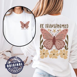 Op de afbeelding: Wit T-shirt met een roze vlinderprint en de tekst "Romeinen 12:2". De achterkant van het shirt heeft een grotere roze vlinderprint met gele bloemen en de tekst "WORD GETRANSFORMEERD".