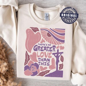 Puede incluir: Una sudadera beige con un diseño gráfico que presenta una cruz, corazones, flores y el texto "There's no greater love than this".