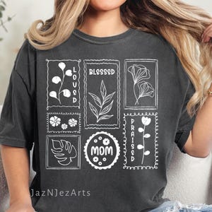 May include: Dark gray t-shirt with white floral and botanical designs, and the words "LOVED", "BLESSED", "PRAISED", and "MOM". The shirt has a vintage, slightly faded look, with the text and graphics in a clean, minimalist style.