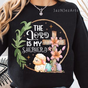 May include: Black sweatshirt featuring a religious design. The design includes a wooden cross, flowers, a lamb, and the text "The Lord is my Shepherd". The design is surrounded by a circular frame and green leaves.