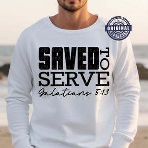 May include: White sweatshirt with the text "SAVED TO SERVE Galatians 5:13" in black. The sweatshirt has a crew neck and long sleeves. A circular "ORIGINAL DESIGNER" logo is in the upper right corner.