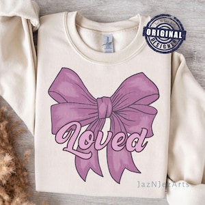 Puede incluir: Una sudadera beige claro con un gran lazo rosa y la palabra "Loved" escrita en rosa.