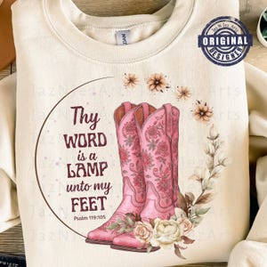 Puede incluir: Sudadera crema con un diseño de botas de vaquero rosas, detalles florales y el texto "Thy WORD is a LAMP unto my FEET" del Salmo 119:105. El diseño incluye un marco circular y flores decorativas.