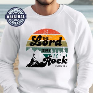 May include: White long-sleeve shirt with a vintage-style graphic of a mountain range, a hiker, and a sunset. The graphic includes the text "The Lord is my Rock" and "Psalm 18:2".