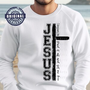 May include: White long-sleeved shirt featuring a black graphic of a cross and the word "JESUS" in a distressed font. The text "Colossians 2:14 Paid it all and set me free" is written in a smaller font next to the graphic.