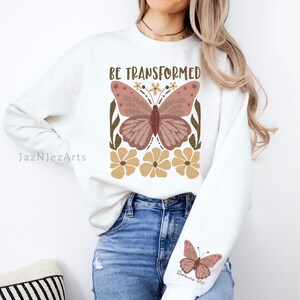 Może przedstawiać: Biała bluza z nadrukiem brązowego motyla i tekstem "BE TRANSFORMED" w brązowych literach. Motyl ma r&oacute;żowe skrzydła i jest otoczony ż&oacute;łtymi kwiatami. Bluza ma r&oacute;wnież mniejszy nadruk motyla na rękawie z tekstem "Rzymian 12:2" w brązowych literach.