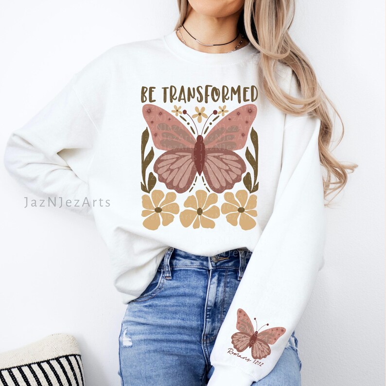 Może przedstawiać: Biała bluza z nadrukiem brązowego motyla i tekstem "BE TRANSFORMED" w brązowych literach. Motyl ma r&oacute;żowe skrzydła i jest otoczony ż&oacute;łtymi kwiatami. Bluza ma r&oacute;wnież mniejszy nadruk motyla na rękawie z tekstem "Rzymian 12:2" w brązowych literach.