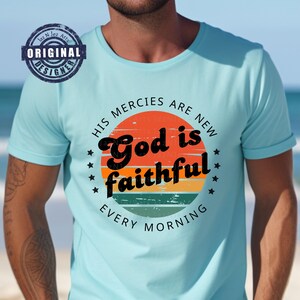 Puede incluir: Una camiseta azul claro con un diseño gráfico de estilo vintage que presenta una salida del sol con el texto "His mercies are new, God is faithful, every morning".