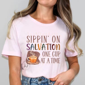 May include: A pink t-shirt with the text "Sippin' on Salvation One Cup at a Time" and a cartoon illustration of a steaming cup of coffee.
