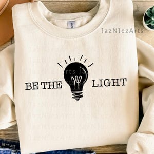 May include: Cream-colored sweatshirt with the phrase "BE THE LIGHT" and a black lightbulb graphic. The design is centered on the front of the long-sleeved top. The brand name "JazNJezArts" is printed on the top right.