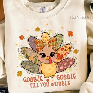 May include: Cream-colored sweatshirt featuring a cartoon turkey with a plaid bow and patterned tail feathers. The turkey has large eyes and a red wattle. The text "GOBBLE TILL YOU WOBBLE" is printed at the bottom. Autumn-themed apparel.