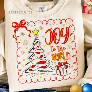 Peut inclure: Sweat-shirt crème avec un motif de Noël festif. Le graphisme présente un sapin de Noël, un cadeau et les mots "JOY to the WORLD" en rouge et jaune. Le motif est souligné d'une bordure rouge et comprend des étoiles et des flocons de neige dorés.