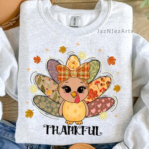 Peut inclure: Sweat-shirt gris clair avec un motif de dinde de dessin animé, un nœud à carreaux et des plumes de queue colorées. La dinde est entourée de feuilles d'automne et de fleurs, avec le mot "THANKFUL" imprimé en dessous. Sweat-shirt de marque Gildan.