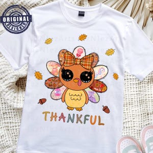 Könnte beinhalten: Weißes T-Shirt mit einem Cartoon-Truthahn-Design, das eine karierte Schleife und bunte Schwanzfedern aufweist. Das Wort "THANKFUL" steht in bunten Buchstaben darunter. Das Shirt hat ein "Original Designer" Logo.