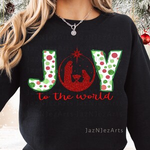 May include: Black sweatshirt with the word "JOY" in green and red polka dot letters. The letter "O" features a red glitter nativity scene. The words "to the world" are in red script below the word "JOY".