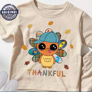 Könnte beinhalten: Beiges Kleinkind-T-Shirt mit einer Cartoon-Truthahnfigur, die eine blaue Kappe trägt. Der Truthahn hat große Augen und bunte Schwanzfedern. Das Wort "THANKFUL" steht in bunten Buchstaben unter dem Truthahn, mit fallenden Blättern und Eicheln.