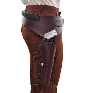 Puede incluir: Un conjunto de funda para pistola y cinturón ancho de cuero marrón oscuro con una tira negra para munición en el borde superior. Una pequeña tarjeta rectangular con el texto Stockhorn Leathers está insertada en la funda, que incluye un broche metálico.