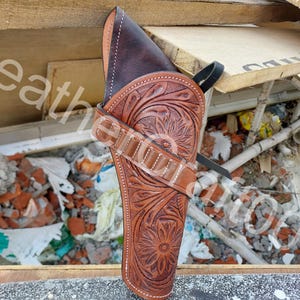 Può includere: Una fondina in pelle marrone con lavorazione floreale e cuciture bianche. La fondina ha un passante per cintura e un cinturino. La pelle è di colore marrone medio e il design floreale è profondamente impresso.
