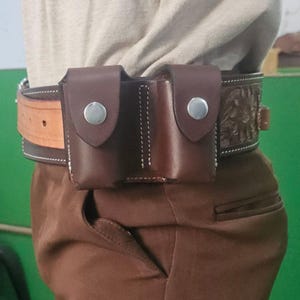 Peut inclure: Une ceinture en cuir marron avec une double pochette attachée. Les pochettes ont des fermetures à pression argentées et des coutures blanches. La ceinture est portée avec un pantalon marron et une chemise de couleur claire. La ceinture a un motif floral.
