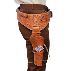 Könnte beinhalten: Ein Pistolenholster und Gürtelset aus hellbraunem Leder mit aufwendigen floralen Prägungen und Mustern. Das Holster enthält ein kleines weißes Etikett mit dem in Schwarz gedruckten Text Deadwood Leather Co. an der Seite des Ledergehäuses.