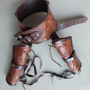 Può includere: Una cintura per pistola in pelle marrone con due fondine abbinate. La cintura e le fondine sono decorate con motivi floreali.