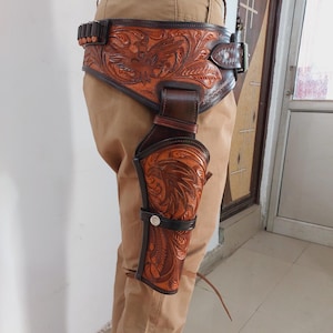 Op de afbeelding: Een bruine leren pistoolholster met ingewikkelde bloemmotieven. De holster is bevestigd aan een bruine leren riem met een zilveren gesp.