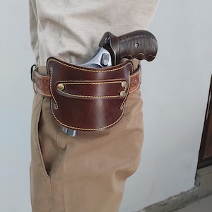 Puede incluir: Una funda de cuero marrón con un revólver. La funda está sujeta a un cinturón marrón con costuras decorativas y remaches dorados. El revólver tiene una empuñadura marrón oscura y un cañón plateado.