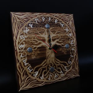 May include: Square wooden clock with a tree of life design. The clock face features a detailed tree carving with runic symbols around the perimeter. The clock hands are black with a red second hand. The clock is approximately 12 inches square.