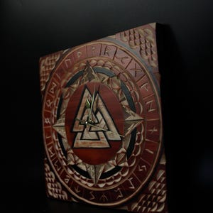 May include: Square wooden wall clock with a carved Viking design. The clock face features a central Valknut symbol, surrounded by runes and geometric patterns. The clock has a dark brown finish with lighter wood accents and gold-colored clock hands.