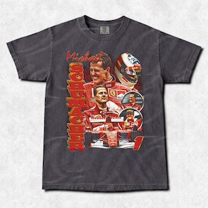 Könnte beinhalten: Dunkelgraues T-Shirt mit einem Design, das mehrere Bilder von Michael Schumacher in seiner Rennkleidung zeigt. Der Text "Michael Schumacher" ist in Rot und Gold gehalten, mit Bildern seines Helms und seines Rennwagens.