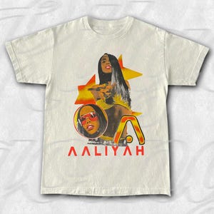 May include: Cream-coloured t-shirt featuring a graphic of Aaliyah. The design includes a portrait, a star, and the word "AALIYAH" in red. The image has a vintage, retro style.