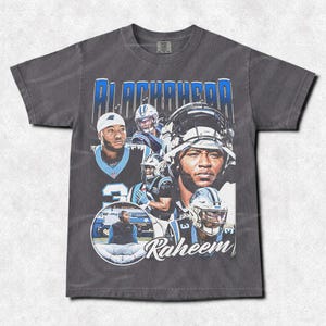 Pode incluir: Camiseta cinza escuro com uma colagem de jogadores de futebol americano em azul e branco. A camiseta tem o nome "BLACKWOOD" em letras azuis e o nome "Raheem" em escrita cursiva, criando um estilo vintage.