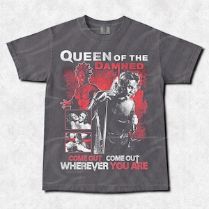 May include: Dark gray t-shirt with a graphic design featuring the text "QUEEN OF THE DAMNED" and the phrases "COME OUT WHEREVER YOU ARE" and "COME OUT". The design includes illustrations in red and black.