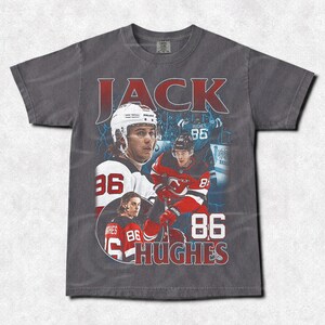 May include: A gray t-shirt featuring a vintage-style design with the name "JACK" in red block letters. The shirt showcases images of a hockey player in a white helmet and red jersey, with the number "86" prominently displayed. The name "HUGHES" is also visible.
