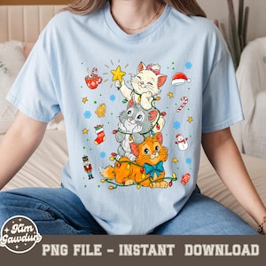 May include: Light blue t-shirt featuring a stack of three cartoon cats decorated with Christmas lights and ornaments. The cats are white, gray, and orange, with a blue bow tie on the bottom cat. The shirt has the text "PNG FILE - INSTANT DOWNLOAD".