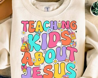 May include: A beige sweatshirt with the text "TEACHING KIDS ABOUT JESUS" in colorful, retro-style letters. The text is surrounded by various school-themed illustrations, including a backpack, pencils, and a ruler.