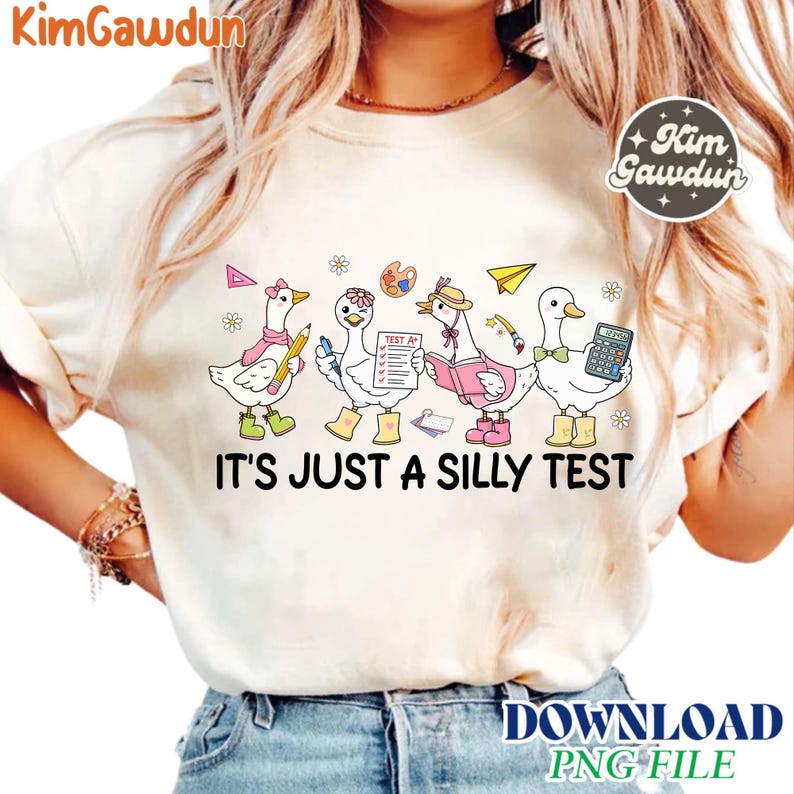 Op de afbeelding: Cr&egrave;mekleurig T-shirt met een grillig ontwerp van cartoon ganzen, elk met school-thema accessoires. De tekst "IT'S JUST A SILLY TEST" staat eronder. Het ontwerp bevat een roze strik, potlood, verfpalet, boek, rekenmachine en papieren vliegtuig.