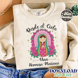 May include: Cream-colored sweatshirt featuring a graphic of a religious figure within a pink halo and floral wreath. The text "Desde el Cielo Una Hermosa Mañana" is displayed in black script. A "Kim Gawdun" logo is visible.