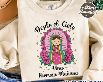 Puede incluir: Sudadera color crema con un gráfico de una figura religiosa dentro de un halo rosa y una corona floral. El texto "Desde el Cielo Una Hermosa Mañana" está en escritura negra. Un logotipo "Kim Gawdun" es visible.