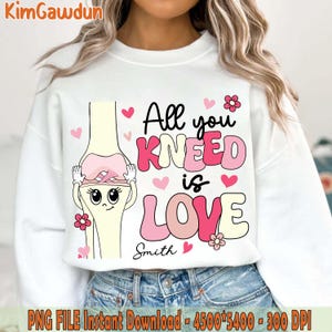 Puede incluir: Sudadera blanca con un dibujo de una articulación de rodilla de dibujos animados que lleva un lazo rosa y el texto "All you kneed is love" con corazones y flores rosas.