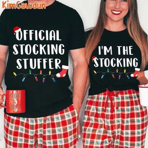 Op de afbeelding: Zwarte T-shirts met kerstthema tekst en afbeeldingen. Eén shirt leest "OFFICIAL STOCKING STUFFER" en de andere "I'M THE STOCKING". Beide shirts hebben een slinger van gekleurde lampjes en een sok-afbeelding. De modellen dragen een rode geruite pyjamabroek.