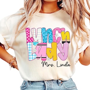 K&ouml;nnte beinhalten: Cremefarbenes T-Shirt mit einem farbenfrohen "Lunch Lady"-Motiv. Der Text besteht aus verschiedenen Mustern, darunter rosa Vichy, blauer Leopardenmuster und gelben Streifen. Der Name "Mrs. Linda" ist darunter gedruckt.