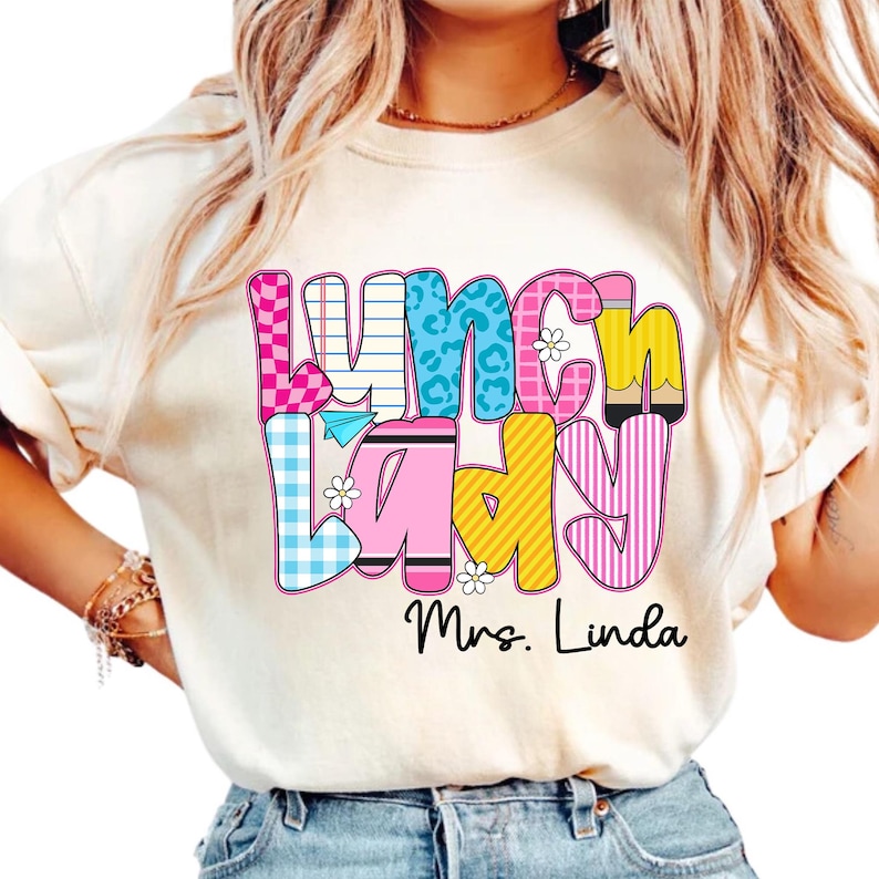K&ouml;nnte beinhalten: Cremefarbenes T-Shirt mit einem farbenfrohen "Lunch Lady"-Motiv. Der Text besteht aus verschiedenen Mustern, darunter rosa Vichy, blauer Leopardenmuster und gelben Streifen. Der Name "Mrs. Linda" ist darunter gedruckt.