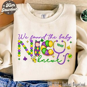 Puede incluir: Una sudadera blanca con un diseño morado, verde y dorado que dice "We found the baby knew". El diseño incluye un bebé en un pañal, una rosca de reyes, un estetoscopio y cuentas de Mardi Gras.