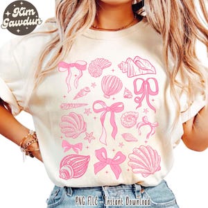 Op de afbeelding: Een wit T-shirt met een roze ontwerp van schelpen, zeesterren en strikken. Het ontwerp wordt herhaald over het hele shirt.