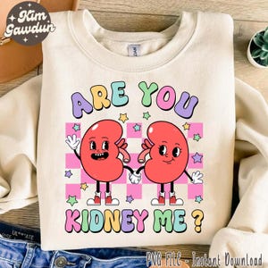 Puede incluir: Sudadera color crema con la frase "ARE YOU KIDNEY ME?" en letras coloridas. El diseño presenta dos riñones de dibujos animados tomados de la mano, sobre un fondo a cuadros rosa y blanco con estrellas.