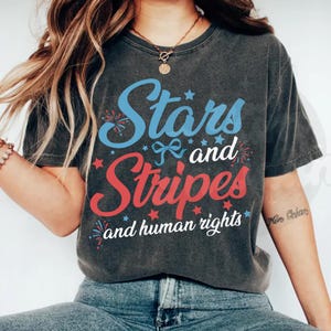 Könnte beinhalten: Ein anthrazitfarbenes Kurzarm-T-Shirt mit dem Text Stars and Stripes and human rights in blauer und roter Schrift. Das Design enthält kleine rote und blaue Sternsymbole, blaue Schleifengrafiken sowie rote und blaue Feuerwerksdarstellungen.