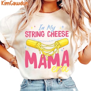 May include: A cream-coloured t-shirt with the text "In My String Cheese Mama Era". The design features a cartoon string cheese with melted cheese strands, and the word "Mama" in pink.