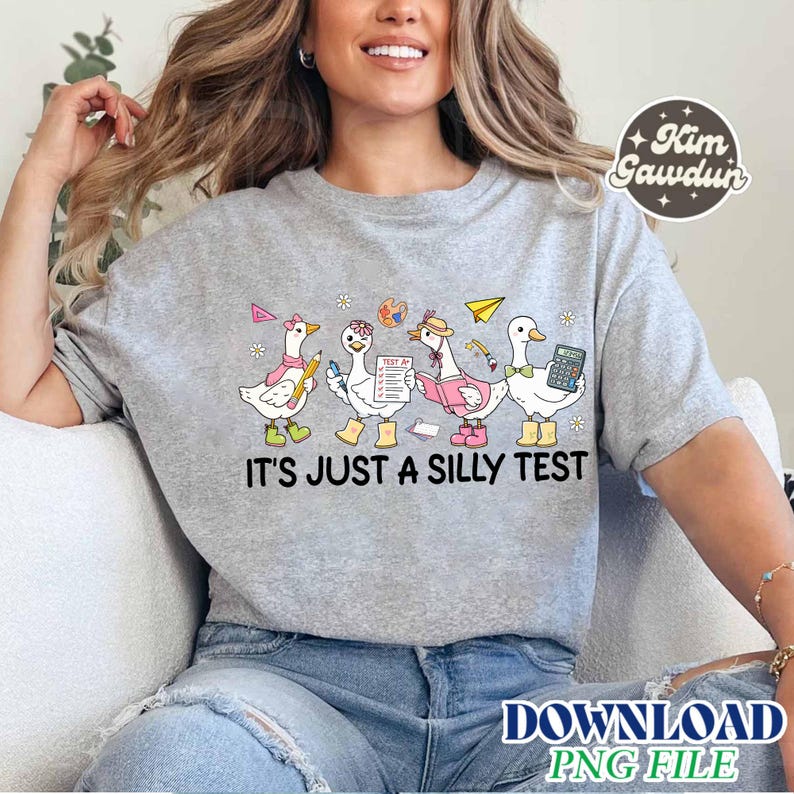 Op de afbeelding: Een lichtgrijs sweatshirt met een grillig ontwerp van vier cartoon ganzen, elk met een uniek accessoire, zoals een potlood, een boek en een rekenmachine. De tekst "IT'S JUST A SILLY TEST" staat eronder gedrukt. Het ontwerp bevat een papieren vliegtuigje en een verfpalet.