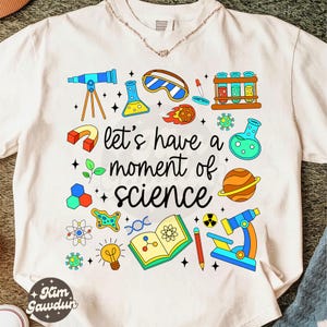 Puede incluir: Camiseta blanca con un gráfico colorido con temática científica. El diseño incluye un telescopio, vasos de precipitados, un microscopio y el texto "let's have a moment of science". La camiseta tiene cuello redondo y mangas cortas.